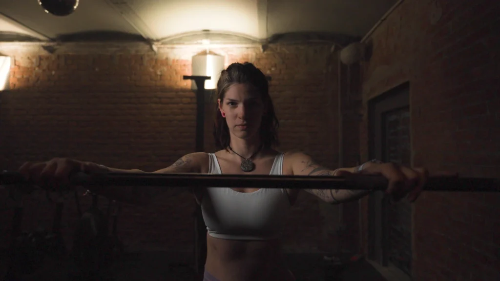 Cinematic Portrait Imagefilm für Fitness Coach Ines Gruber von ArsMotion. Sie hält eine Langhantel mit fokussiertem Blick in einem Gym mit Ziegelwand. Das Licht ist dramatisch und stimmungsvoll.