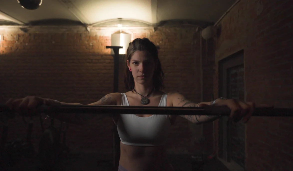 Cinematic Portrait Imagefilm für Fitness Coach Ines Gruber von ArsMotion. Sie hält eine Langhantel mit fokussiertem Blick in einem Gym mit Ziegelwand. Das Licht ist dramatisch und stimmungsvoll.