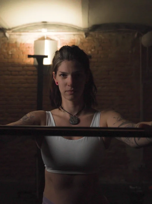 Cinematic Portrait Imagefilm für Fitness Coach Ines Gruber von ArsMotion. Sie hält eine Langhantel mit fokussiertem Blick in einem Gym mit Ziegelwand. Das Licht ist dramatisch und stimmungsvoll.