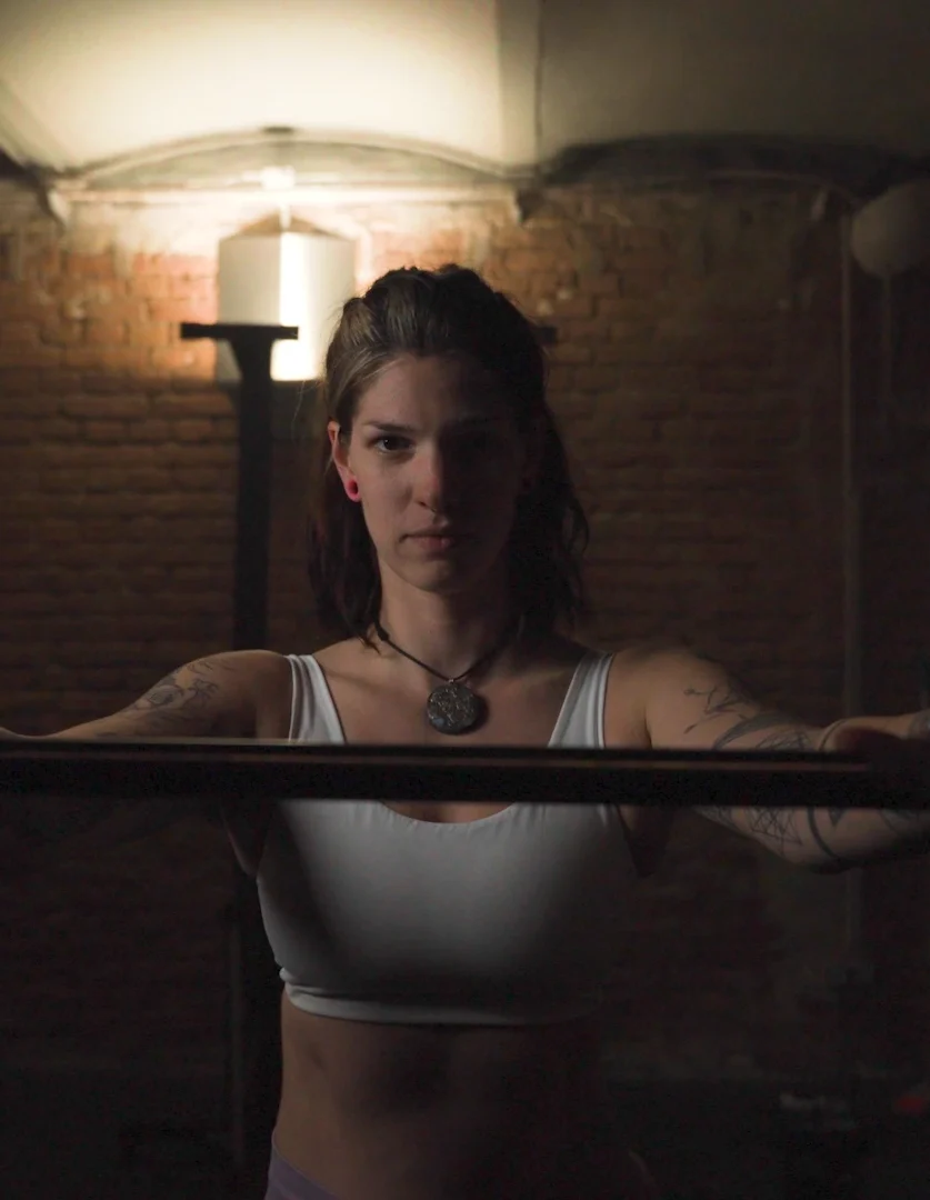 Cinematic Portrait Imagefilm für Fitness Coach Ines Gruber von ArsMotion. Sie hält eine Langhantel mit fokussiertem Blick in einem Gym mit Ziegelwand. Das Licht ist dramatisch und stimmungsvoll.