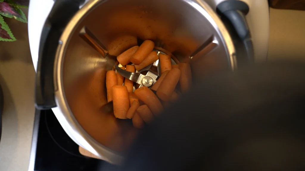 Technische Nahaufnahme in den Edelstahl-Mixtopf des Thermomix: Ganze Karotten liegen bereit zum Zerkleinern über dem Mixmesser – Detailshot für ein Koch-Produktvideo.