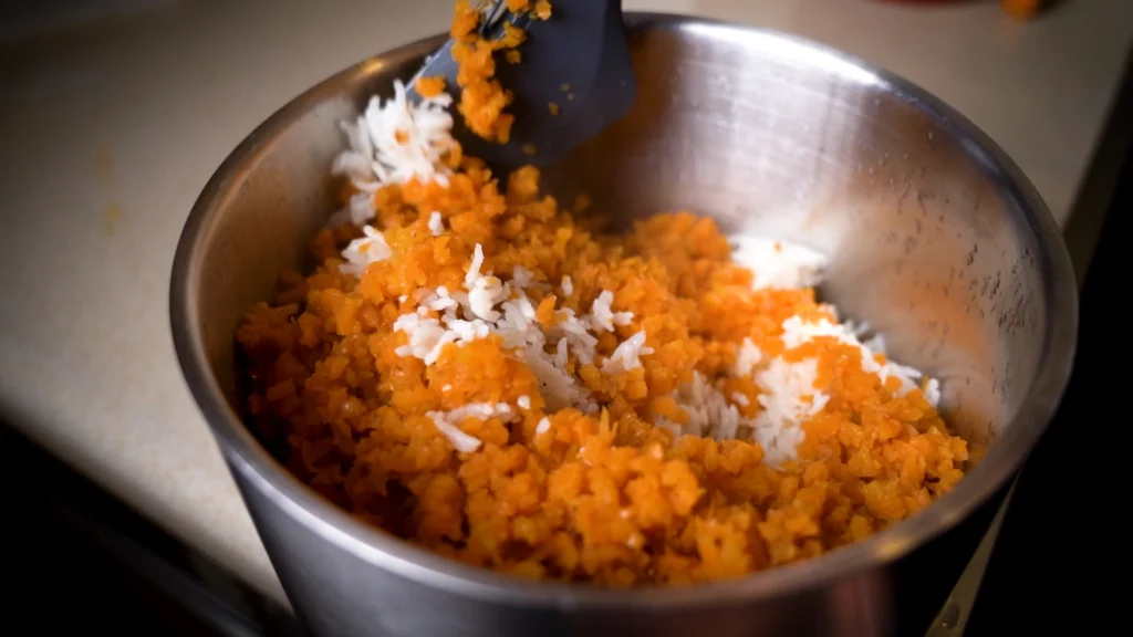 Nahaufnahme aus dem Edelstahl-Mixtopf des Thermomix: Frische, zerkleinerte Karotten und Reis werden mit dem Spatel vermengt.