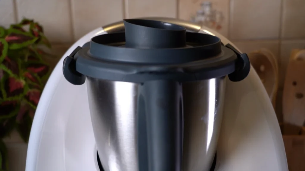 Detailaufnahme des geschlossenen Thermomix-Deckels mit Messbecher während des Kochvorgangs: Leichter Dampf steigt auf und visualisiert die Hitzeentwicklung.