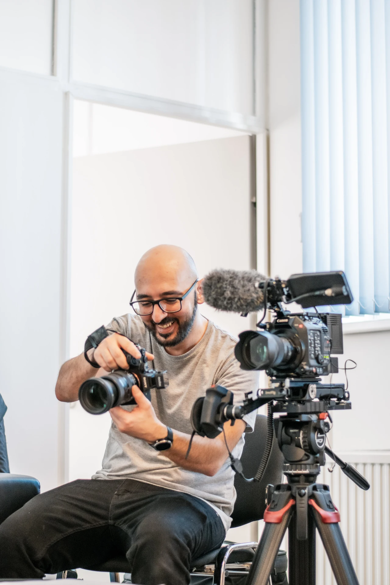 Mediengestalter Arsen Stepanian sitzt lächelnd mit einer Kamera in den Händen an einem Set im Büro. Neben ihm steht eine größere Videokamera mit Mikrofon auf einem Stativ.