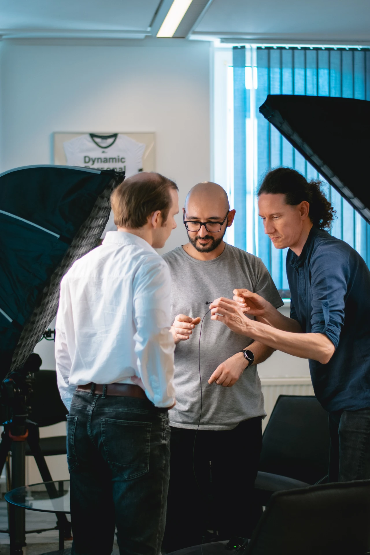Drei Männer in einem Büro, die zusammenarbeiten. Ein Mann in einem grauen T-Shirt (Arsen) hält ein technisches Gerät, während ein Mann in einem weißen Hemd und ein Mann in einem blauen Hemd ihn beobachten.